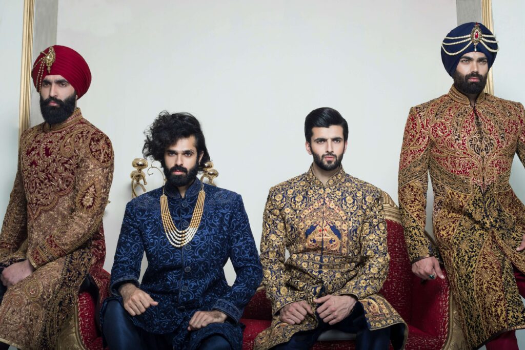 Four grooms showcasing diverse sherwani styles - burgundy embroidered with red turban, navy blue with gold jewelry, golden with intricate patterns, and gold with navy turban and traditional accessories in elegant setting