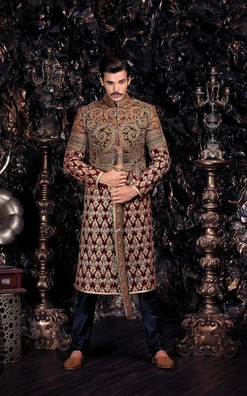 Groom in burgundy and gold embroidered sherwani with traditional high collar standing among ornate brass candelabras in luxurious dark setting